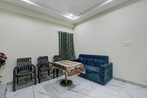 A room with a blue sofa, two chairs, a table with a patterned tablecloth, a green curtain, and a white wall with a ceiling light. - Hotel O RR GRAND STAY (Hyderabad)