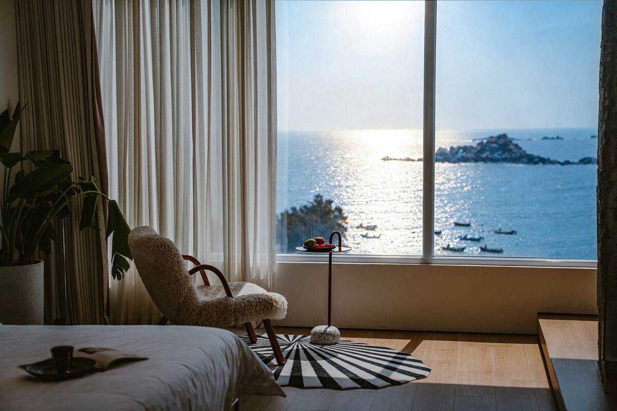 Ein Zimmer mit einem Bett, einem Sessel, einer Topfpflanze, einem Tisch mit Obstschale und Meerblick durch ein großes Fenster.