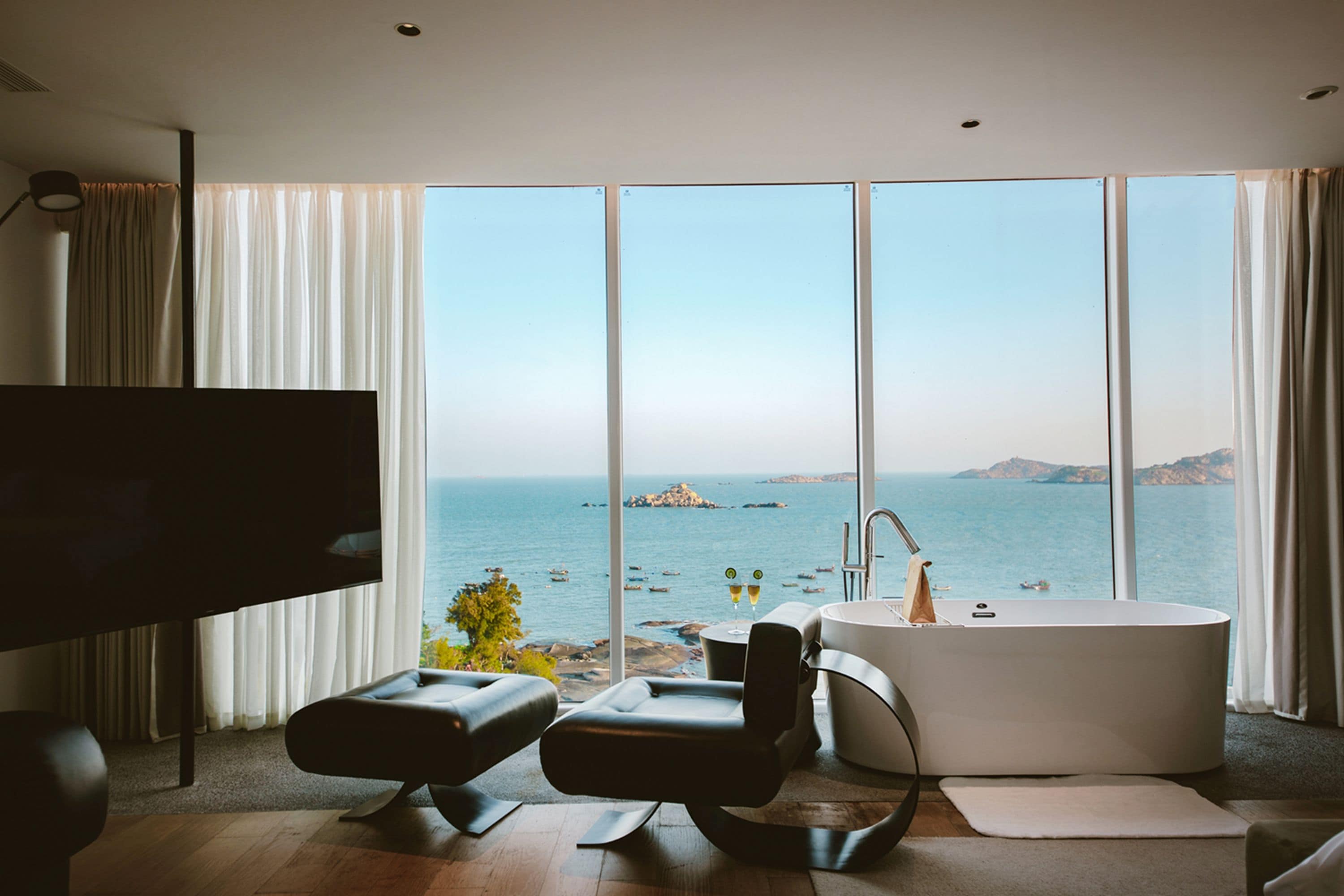Uma sala de estar moderna com uma janela ampla, televisão de tela plana, sofá de couro preto, banheira redonda e piso de madeira.