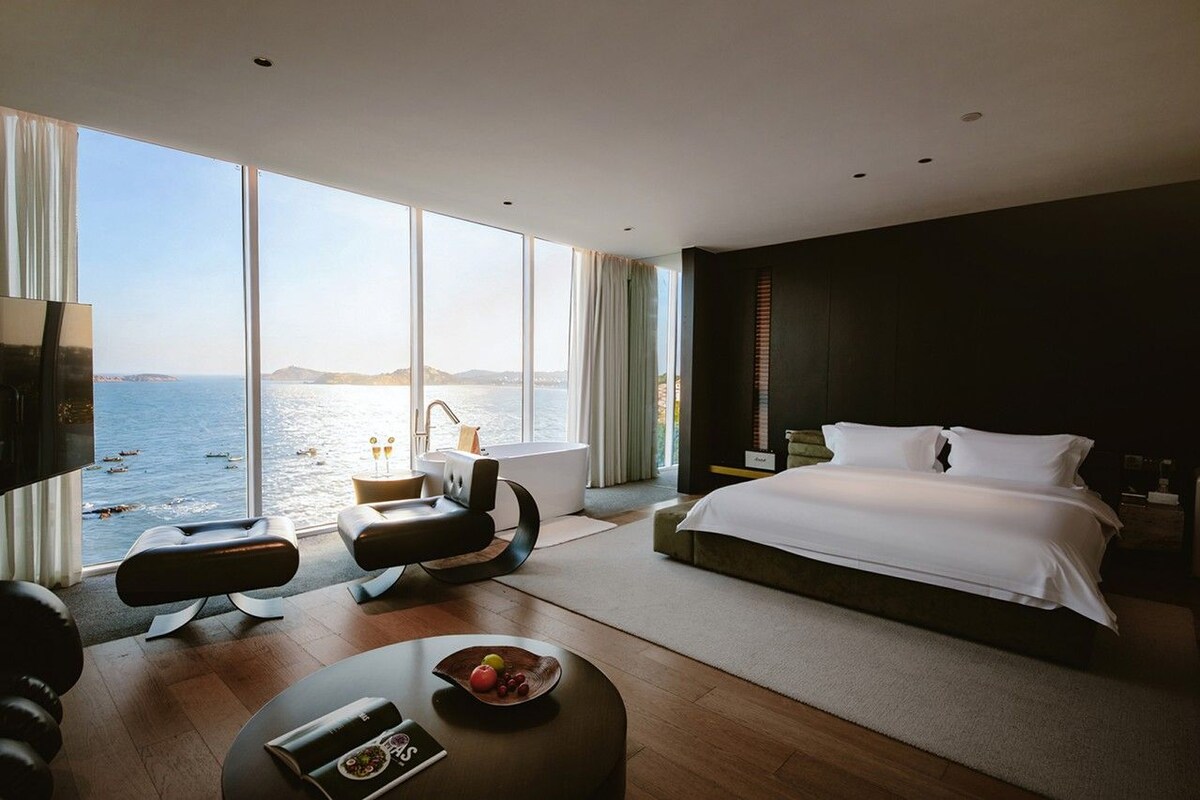 Ein modernes Hotelzimmer mit großem Fenster, einem Bett, einer Sitzecke und einem Esstisch.