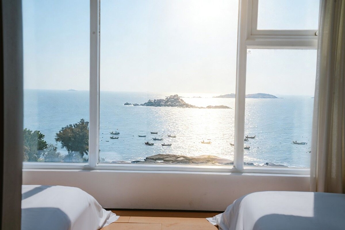 Ein Zimmer mit großem Fenster, das einen ruhigen Meeresblick auf Boote, eine entfernte Insel und einen klaren Himmel bietet.
