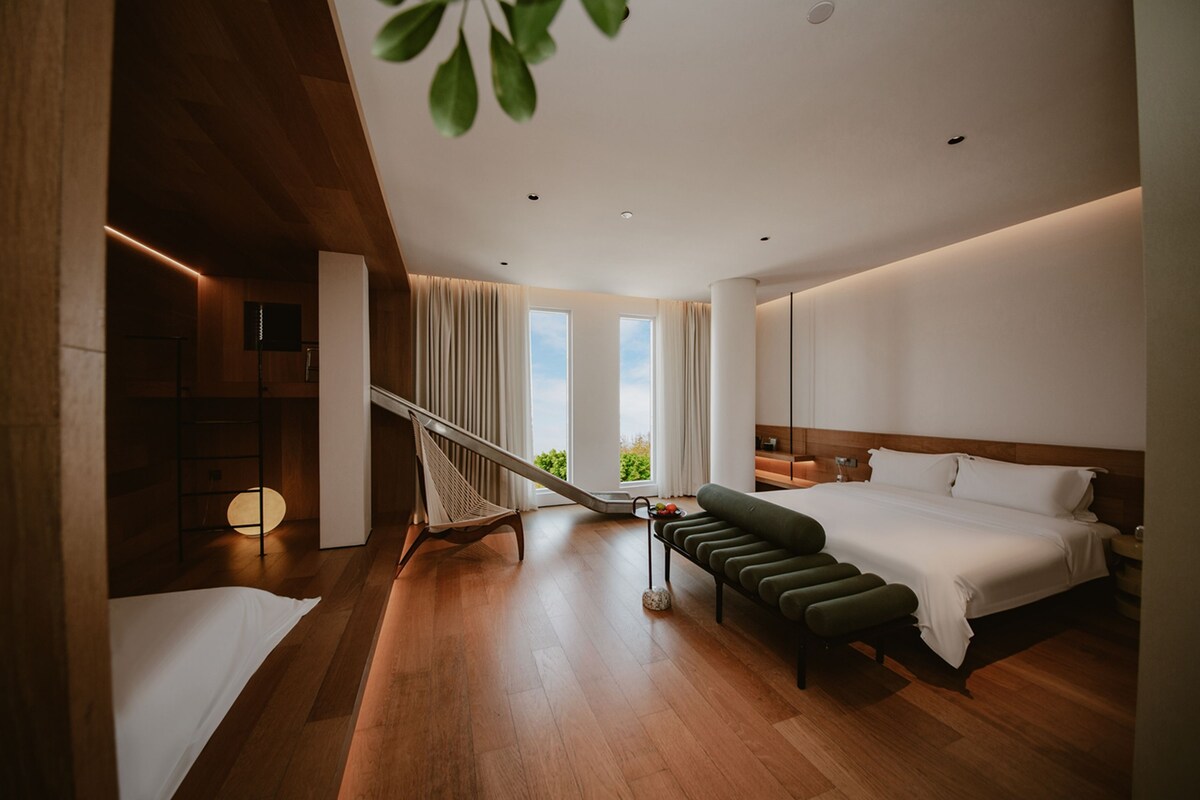 Ein modernes Hotelzimmer mit Holzboden, einem Bett, einer Bank, einer Treppe und Blick auf Grünfläche draußen.