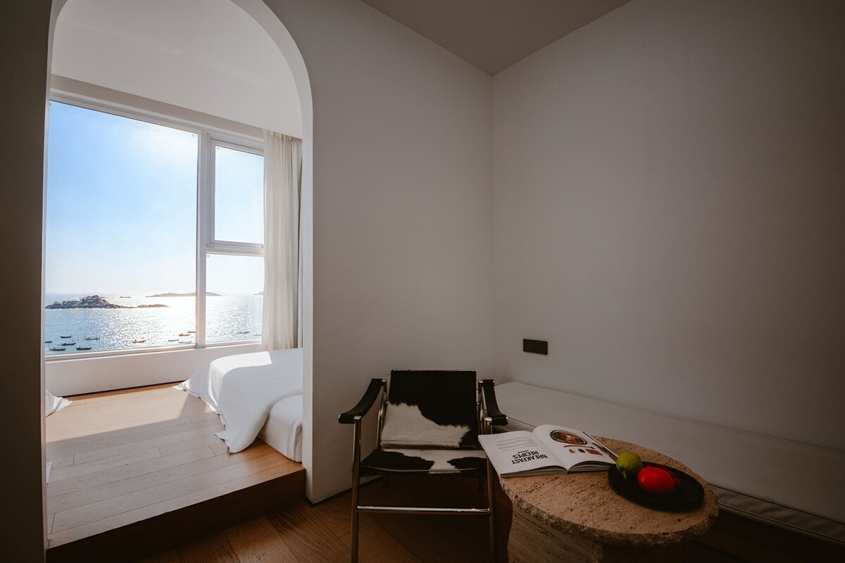 Ein Zimmer mit großem Fenster und Blick auf ein Gewässer, ein Bett, ein Sessel und ein kleiner Tisch mit Buch und Obst.