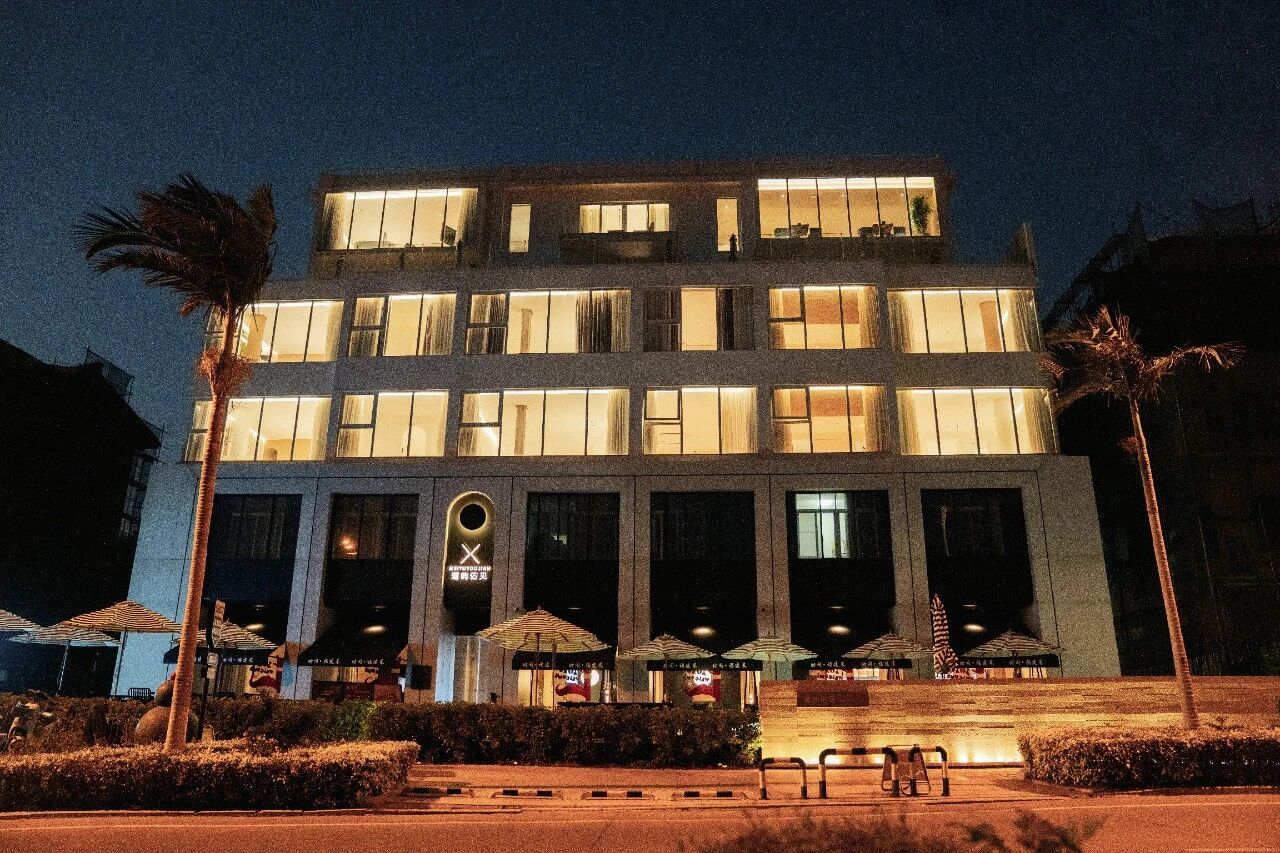 Um hotel com vários andares, janelas iluminadas, entrada central com uma placa e área externa com mesas e guarda-sóis.