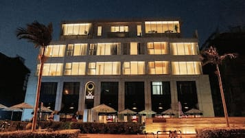 Um hotel com vários andares, janelas iluminadas, entrada central com uma placa e área externa com mesas e guarda-sóis.