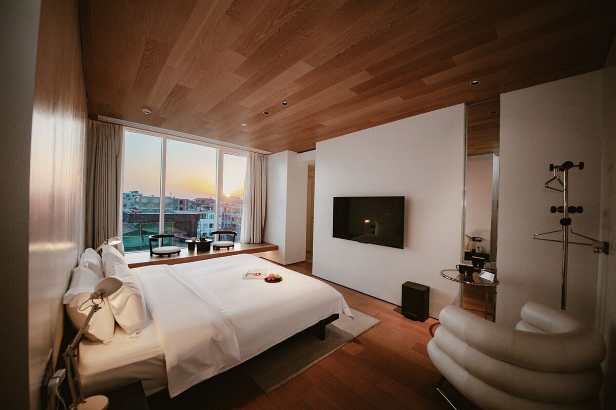 Ein modernes Hotelzimmer mit einem großen Bett, einem Fernseher, Blick auf die Stadt und einer Holzbalkendecke.