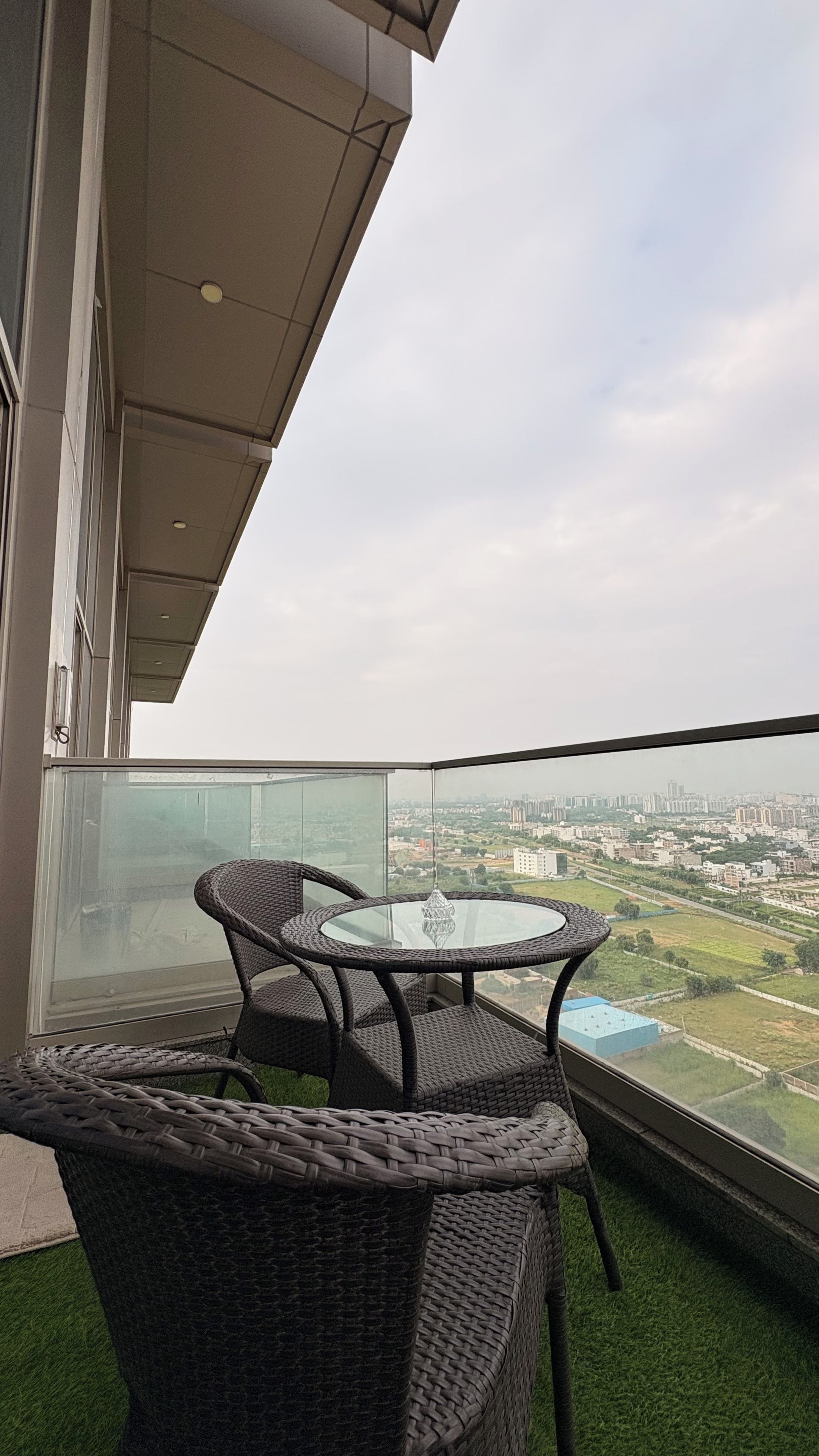 A balcony with wicker furniture and a glass table, overlooking a cityscape.