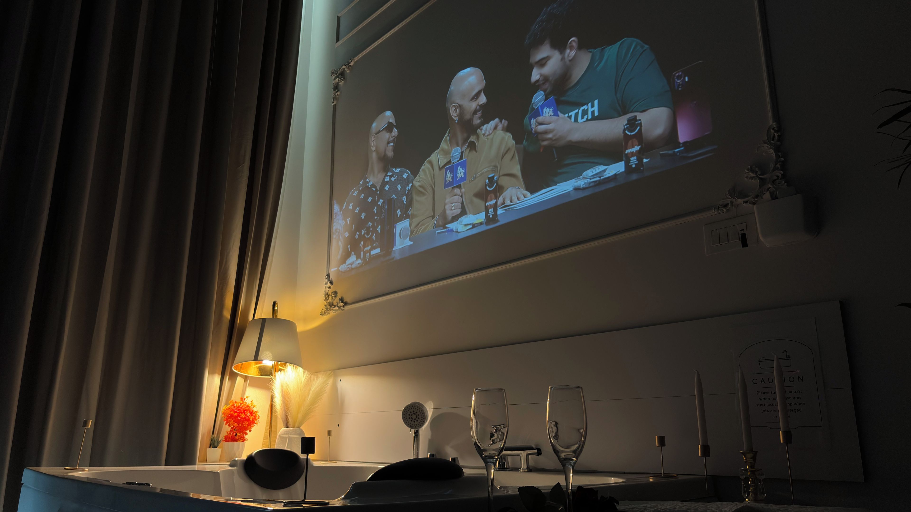 A room with a television screen displaying a scene, a table with a lamp, a vase with flowers, and a candle holder.
