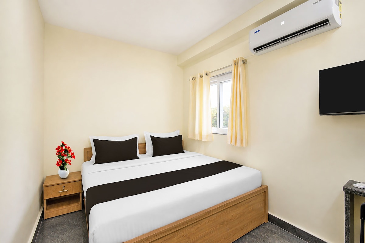 A hotel room with a bed, a nightstand, a vase with flowers, a window with curtains, and a flat-screen TV mounted on the wall.