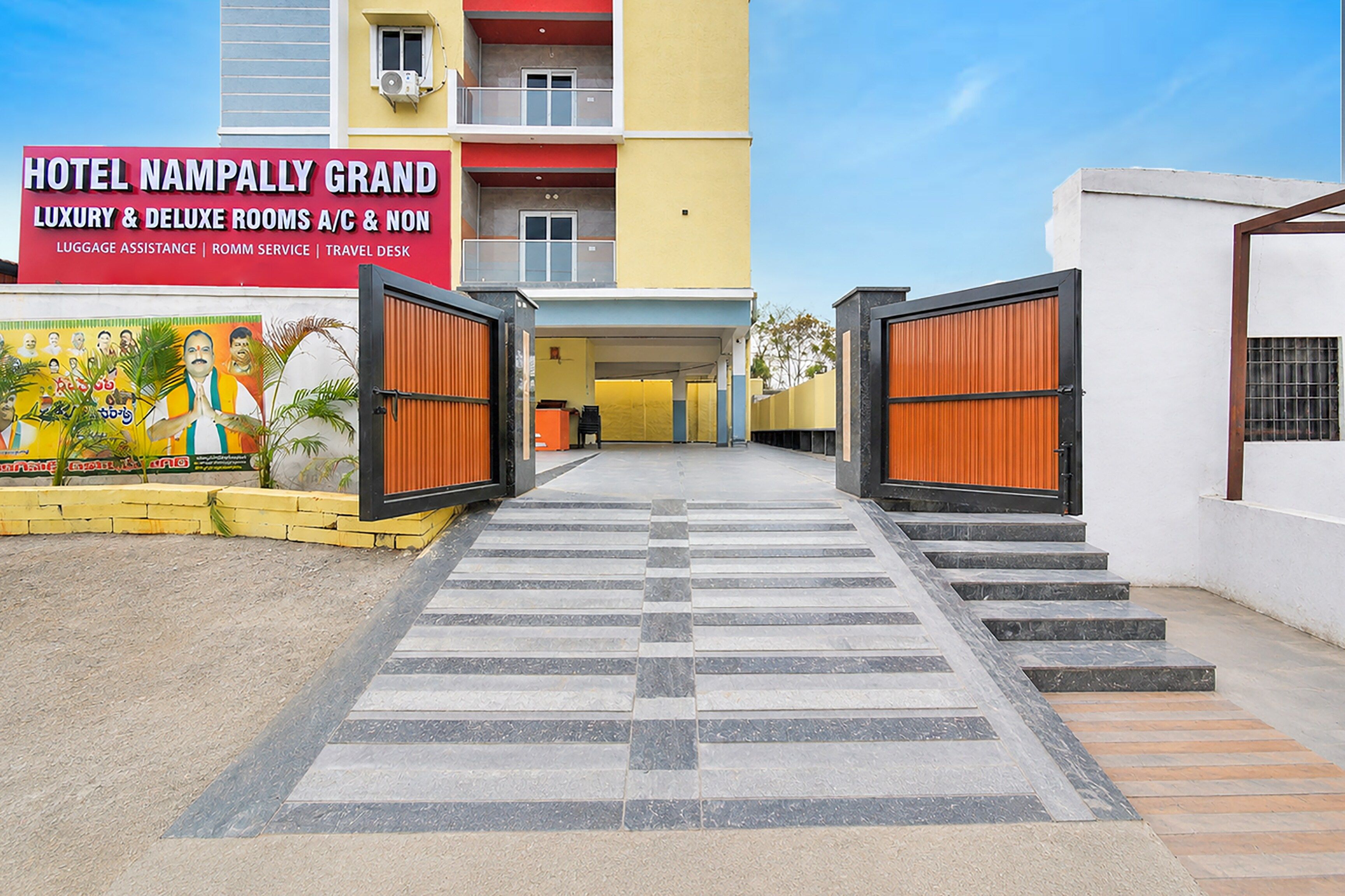 A hotel entrance with a sign for 'HOTEL NAMPALLY GRAND' offering luxury and deluxe rooms, luggage assistance, room service, and a travel desk.