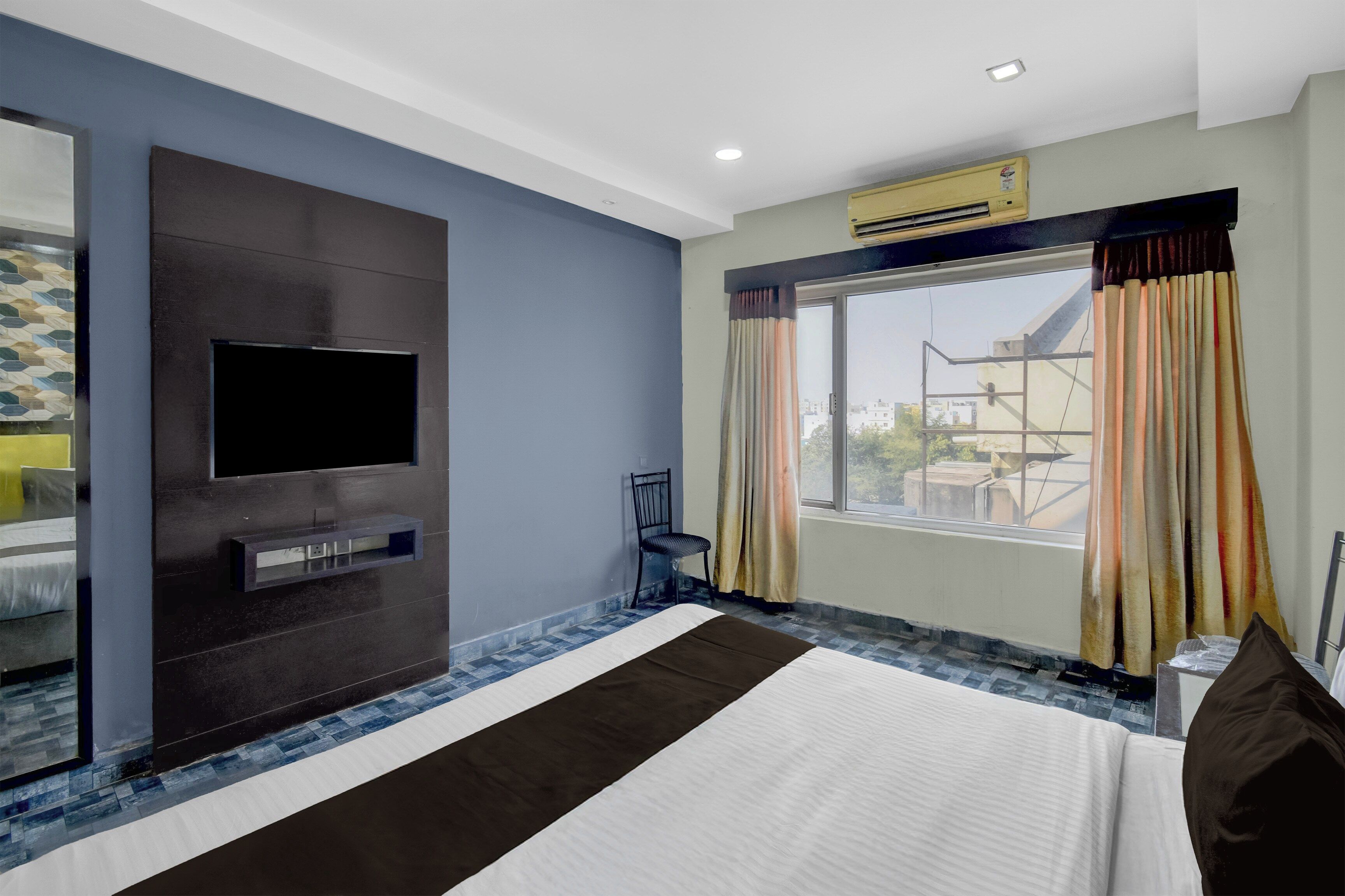 Une chambre d’hôtel dotée d’un téléviseur à écran plat fixé au mur, d’un lit recouvert d’une courtepointe blanche et marron, d’une fenêtre avec des rideaux et offrant une vue sur les bâtiments environnants.