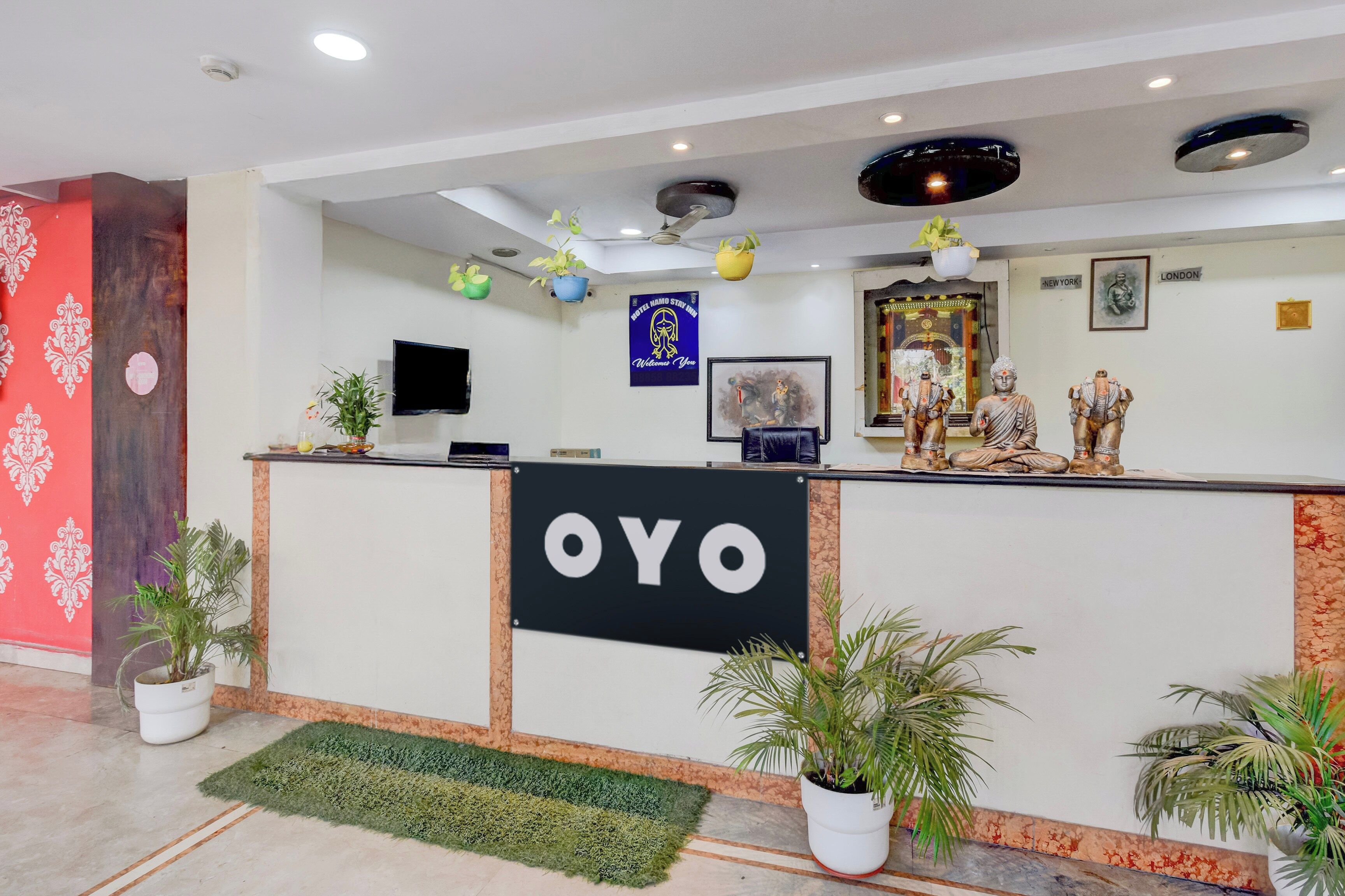 Una recepción de hotel con un cartel que dice 'OYO', un televisor de pantalla plana, un letrero de bienvenida enmarcado y una estatua de Buda.