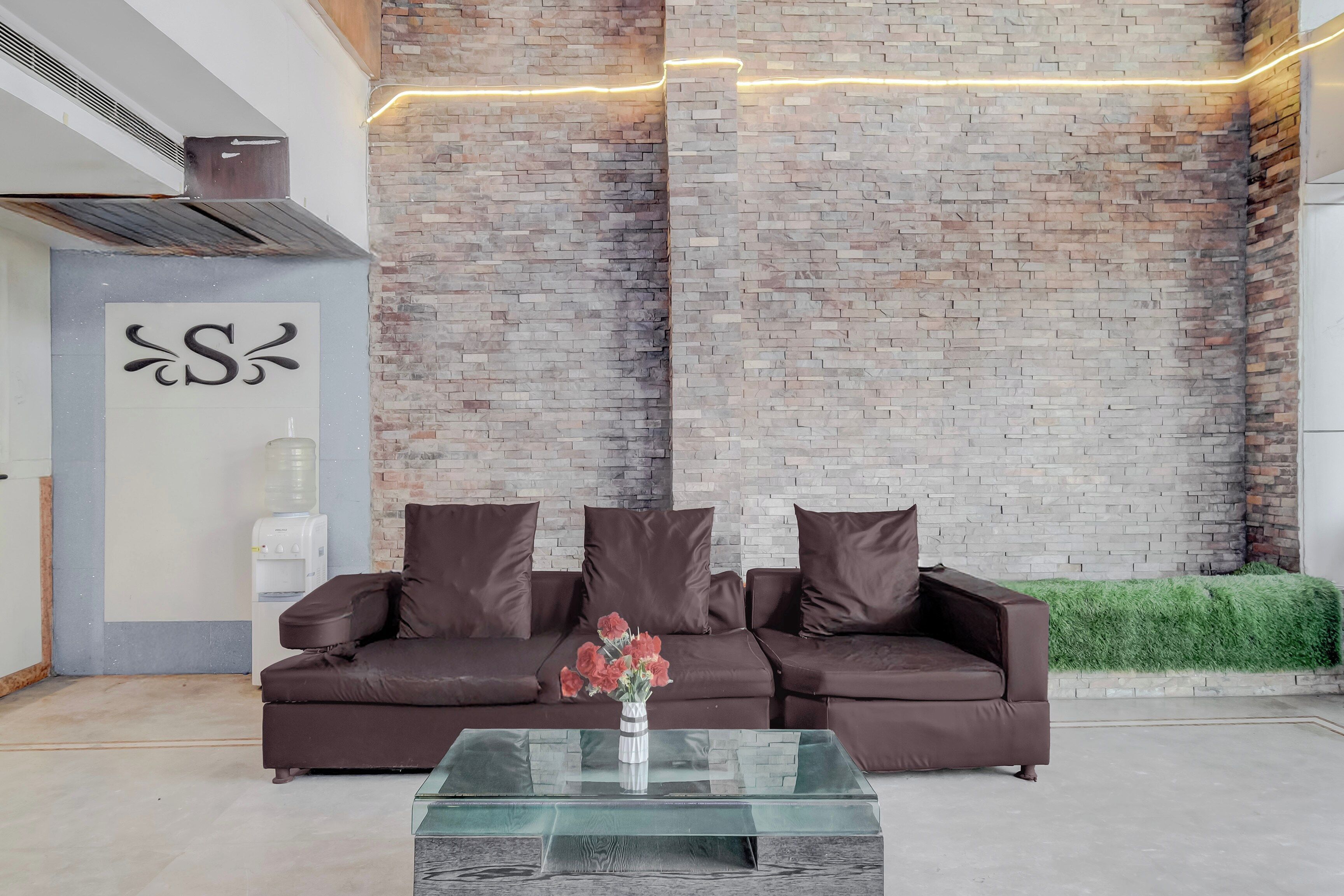 Un espace d’accueil moderne avec un mur en briques, un canapé avec des coussins, une table basse en verre et un distributeur d’eau.