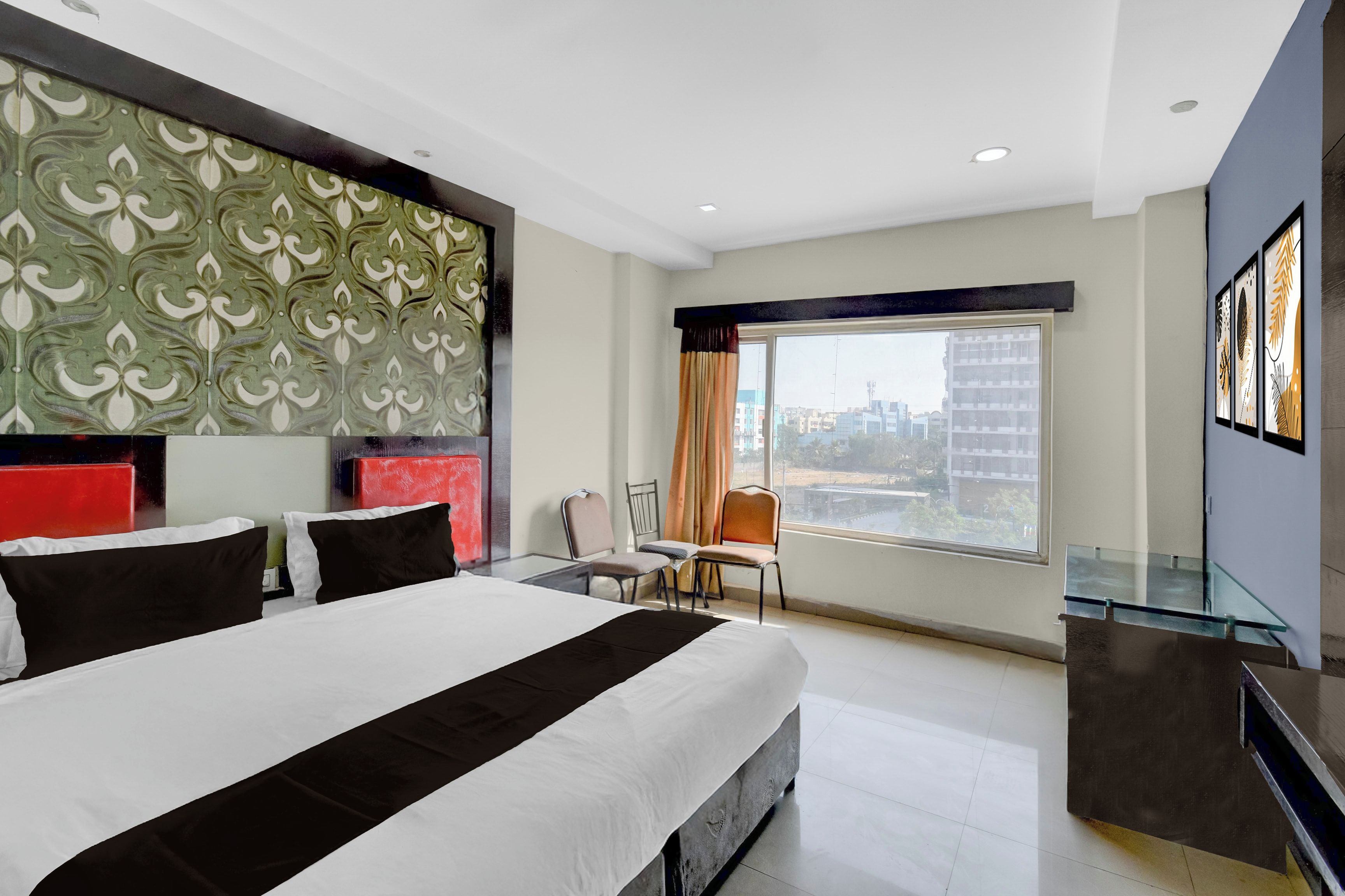 Habitación de hotel con una cama grande, una mesa de vidrio, una silla, un escritorio y vistas a edificios a través de la ventana.