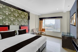 Habitación de hotel con una cama grande, una mesa de vidrio, una silla, un escritorio y vistas a edificios a través de la ventana.