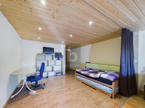 A room with a wooden ceiling, a bed with a patterned bedspread, a desk with a chair, and a shelving unit. - Charming 2-bedroom apartment in Leukerbad perfect for your getaway (Leukerbad)