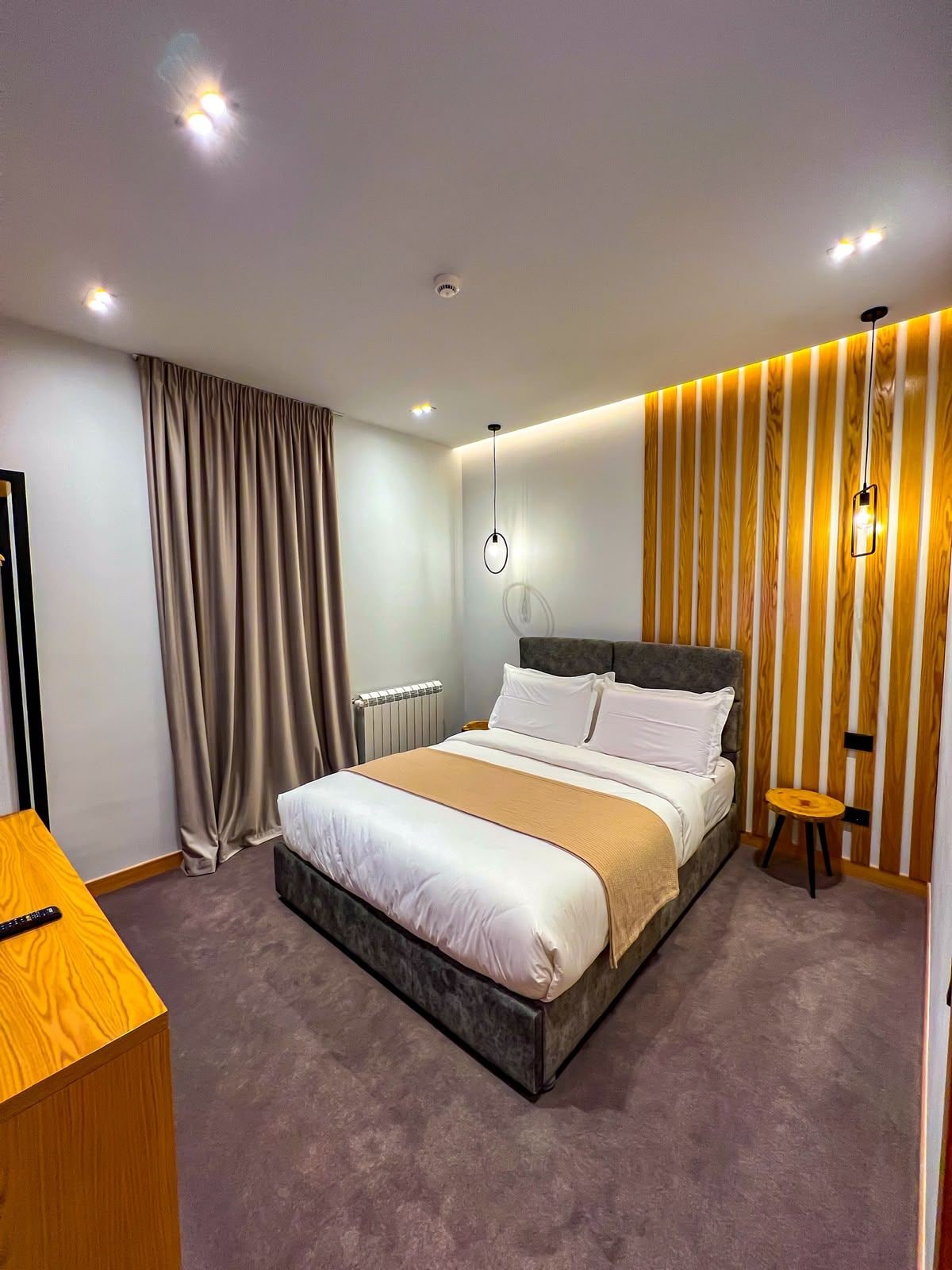 Habitación de hotel moderna con una cama grande, una mesita de noche, un taburete pequeño y una lámpara fijada a la pared.