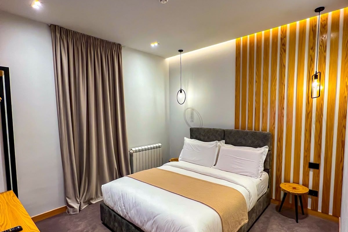 Quarto de hotel moderno com uma cama grande, uma mesinha de cabeceira, um banquinho pequeno e um candeeiro fixado na parede.