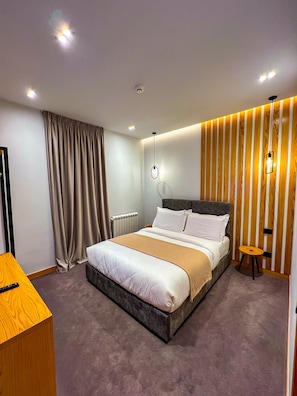 Quarto de hotel moderno com uma cama grande, uma mesinha de cabeceira, um banquinho pequeno e um candeeiro fixado na parede.