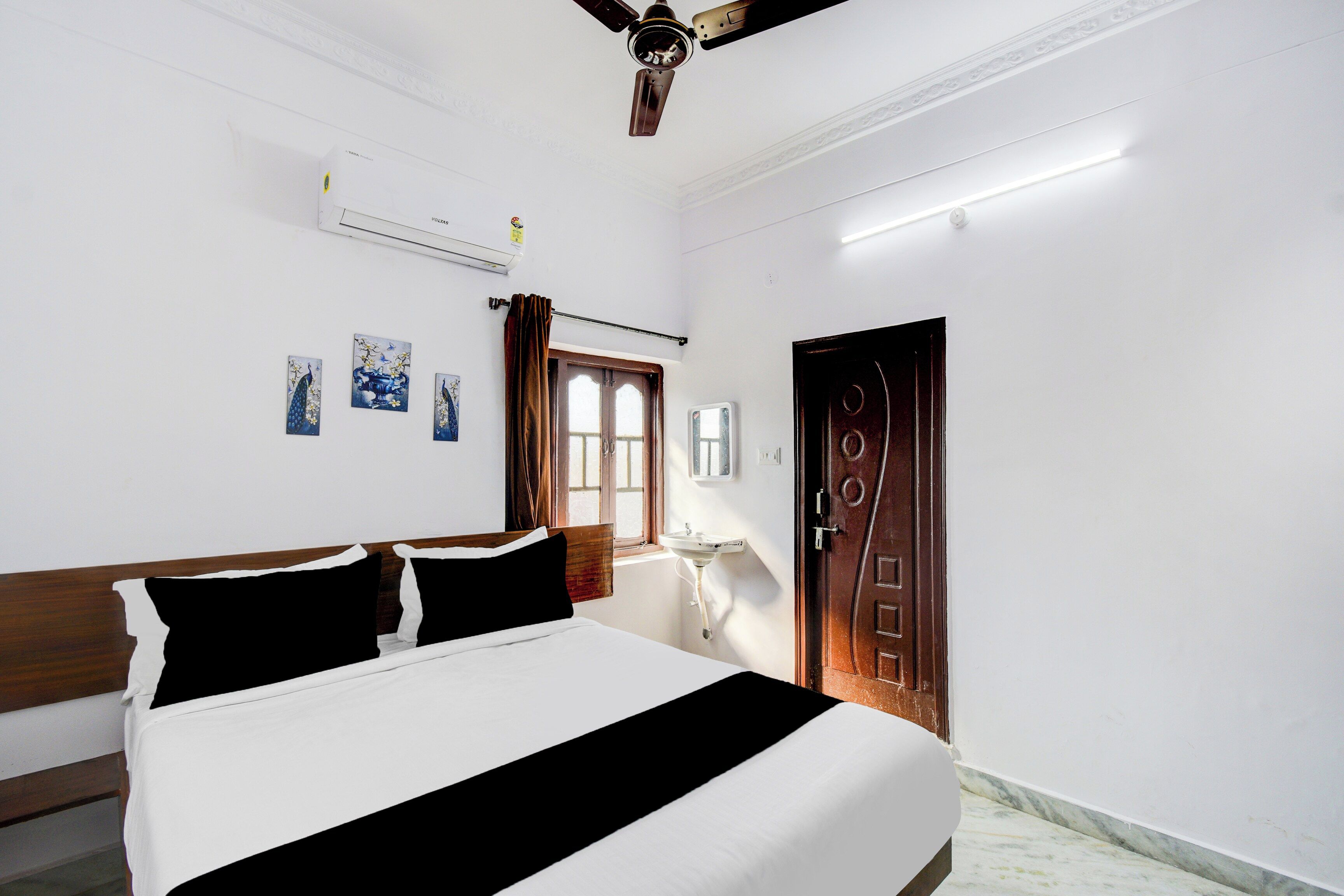 A hotel room with a bed, a ceiling fan, an air conditioning unit, a window with curtains, a door, and wall-mounted artwork.