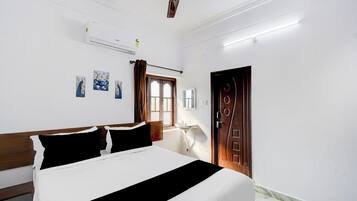 A hotel room with a bed, a ceiling fan, an air conditioning unit, a window with curtains, a door, and wall-mounted artwork.