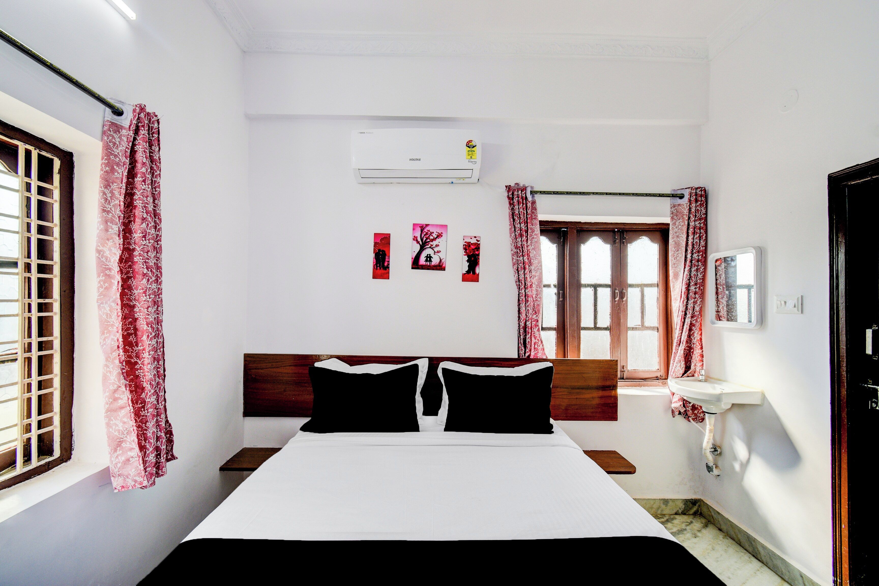 A bedroom with a bed, a window with curtains, a wall-mounted air conditioner, and a small bedside table with a mirror.