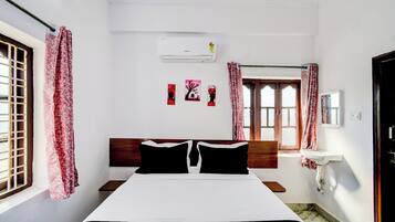 A bedroom with a bed, a window with curtains, a wall-mounted air conditioner, and a small bedside table with a mirror.