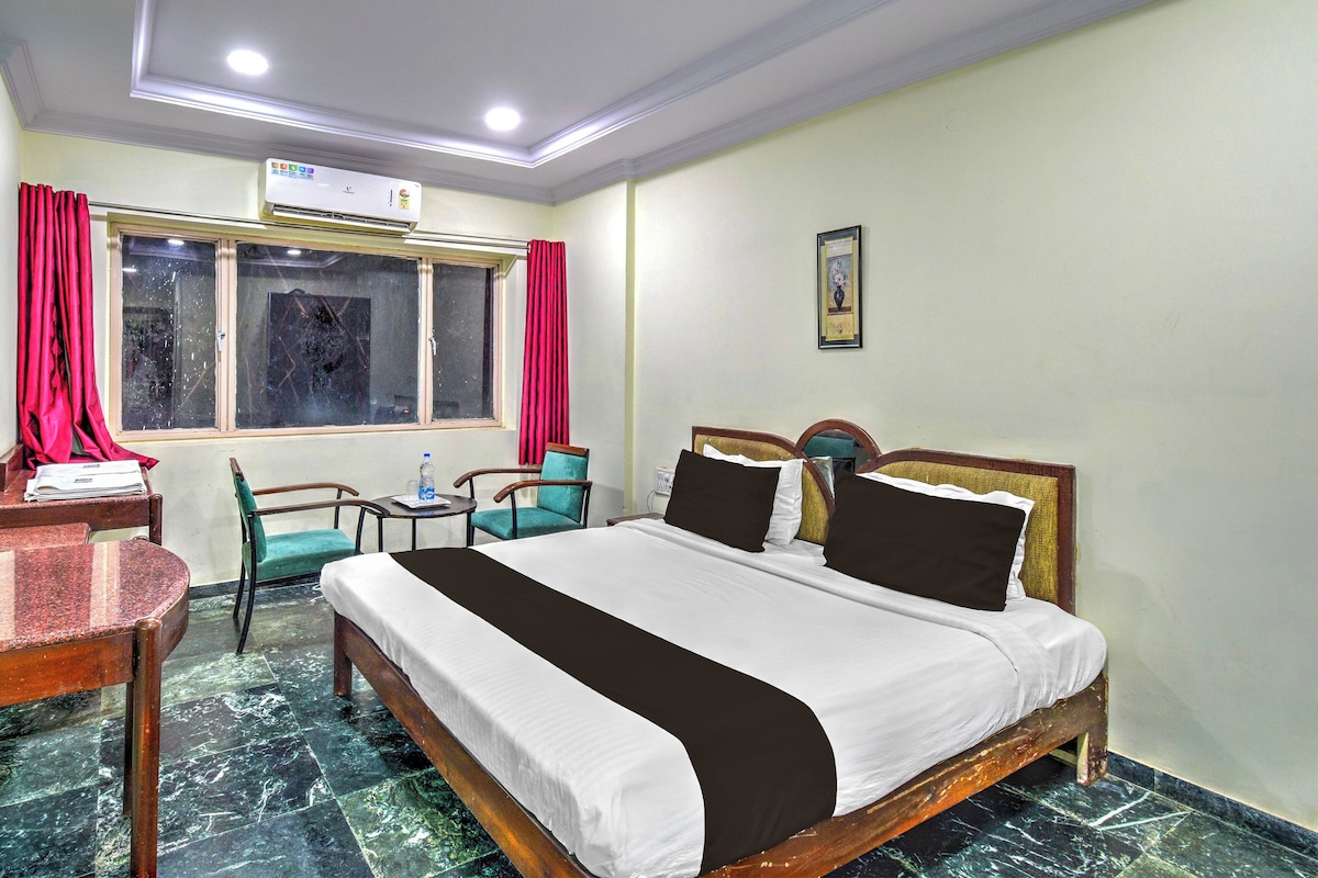 A hotel room with a bed, a desk, a chair, a window with curtains, and a wall-mounted air conditioner.