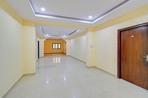 A spacious hallway with a tiled floor, a wooden door on the right, and a window with a black frame at the end. - Hotel O VSM Suites (Hyderabad)