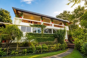 A two-story house with a balcony, surrounded by lush greenery and a stone pathway. - Hotel R.J. House (Aburaj)