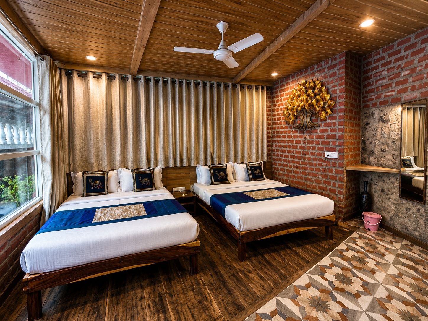 A hotel room with two beds, wooden flooring, a brick accent wall, a ceiling fan, and a window with curtains.