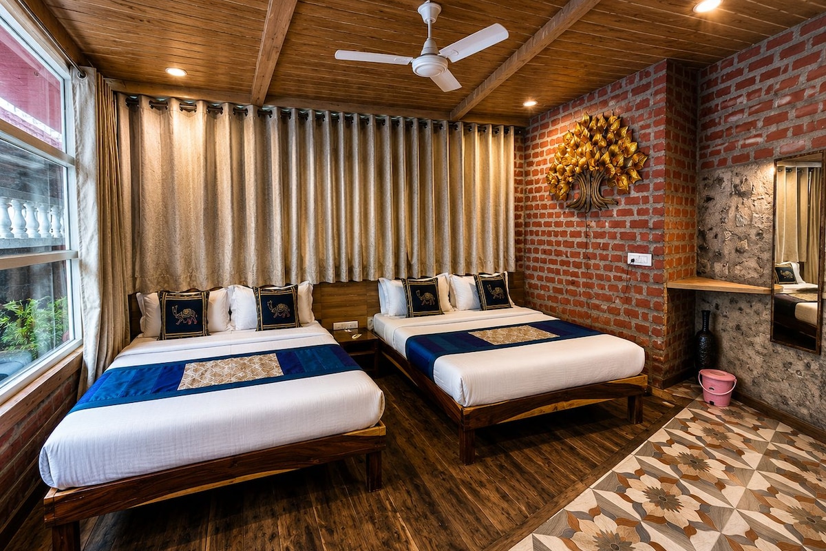 A hotel room with two beds, wooden flooring, a brick accent wall, a ceiling fan, and a window with curtains.