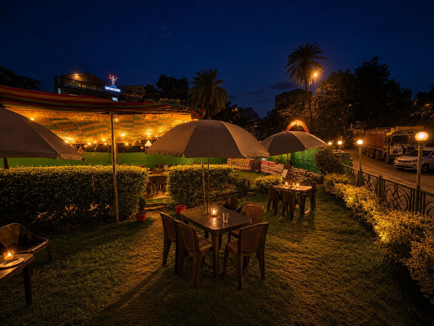 An outdoor dining area with tables and chairs under umbrellas, surrounded by greenery and a road with vehicles.
