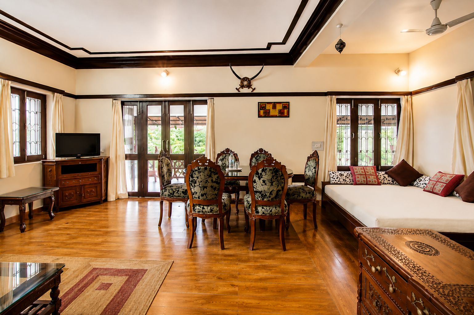 A hotel room with a wooden floor, a bed, a TV, a dining table with chairs, and a ceiling fan.