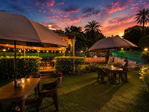 An outdoor seating area with tables and chairs under umbrellas, surrounded by greenery and a sunset sky. - Hotel R.J. House (Aburaj)