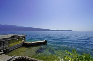 A clear view of a waterfront with a concrete dock extending into calm blue water, surrounded by a clear sky and distant mountains.