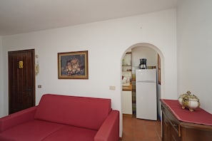 A living room with a red sofa, a framed painting, a wooden door, and a kitchen area with a refrigerator and coffee maker. - Appartamento Bouganville (Toscolano Maderno)