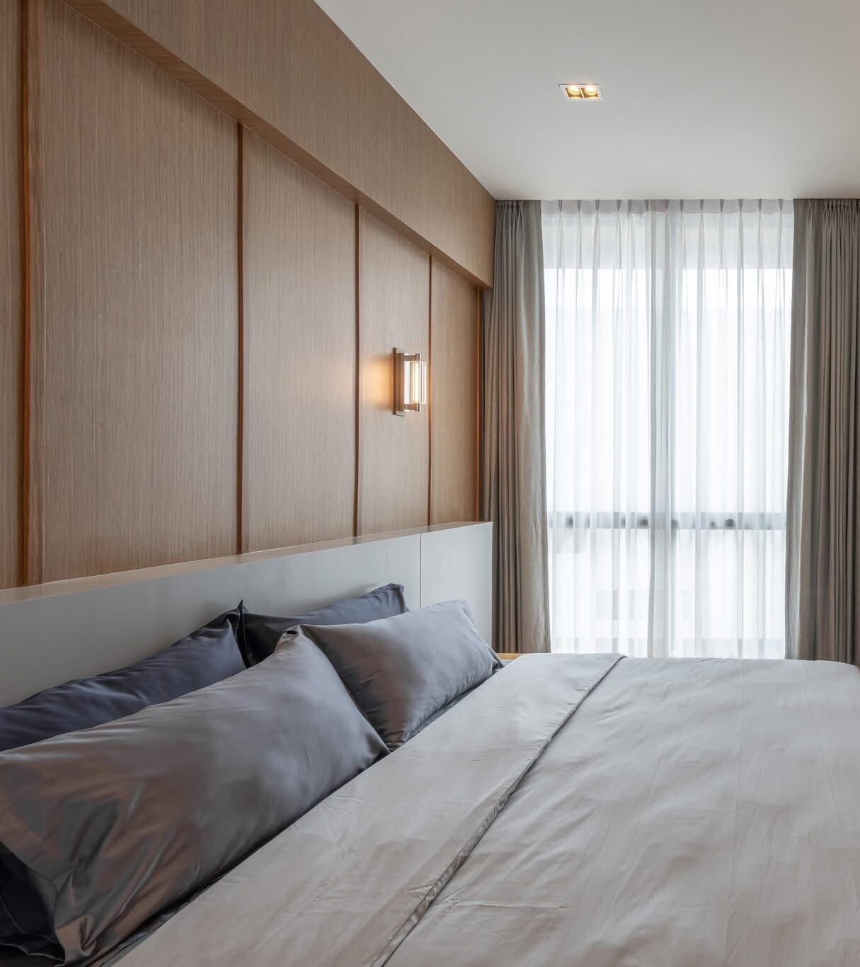 A hotel room with a large bed, a headboard with built-in lighting, and a view through sheer curtains.