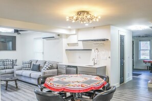 Ein modernes Wohnzimmer mit einem grauen Sofa, einem Esstisch mit blumigem Tischläufer und einer Küchenzeile mit Einbauofen und Spüle.