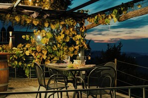 En uteservering med ett bord för två personer, omgiven av stolar, under en pergola som är täckt av grönska.