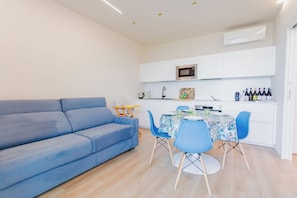 Een moderne woonkamer met een blauwe bank, een eettafel met stoelen, en een keuken met witte kasten en apparatuur.