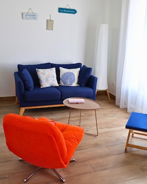 Ein Wohnzimmer mit einem blauen Sofa, zwei Kissen, einem kleinen runden Tisch und einem leuchtend orangefarbenen Sessel.