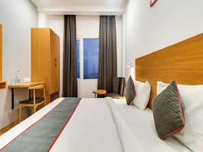 A hotel room with a bed, a desk, a chair, a small table, and a window with curtains. - Townhouse Jashn (Lucknow)