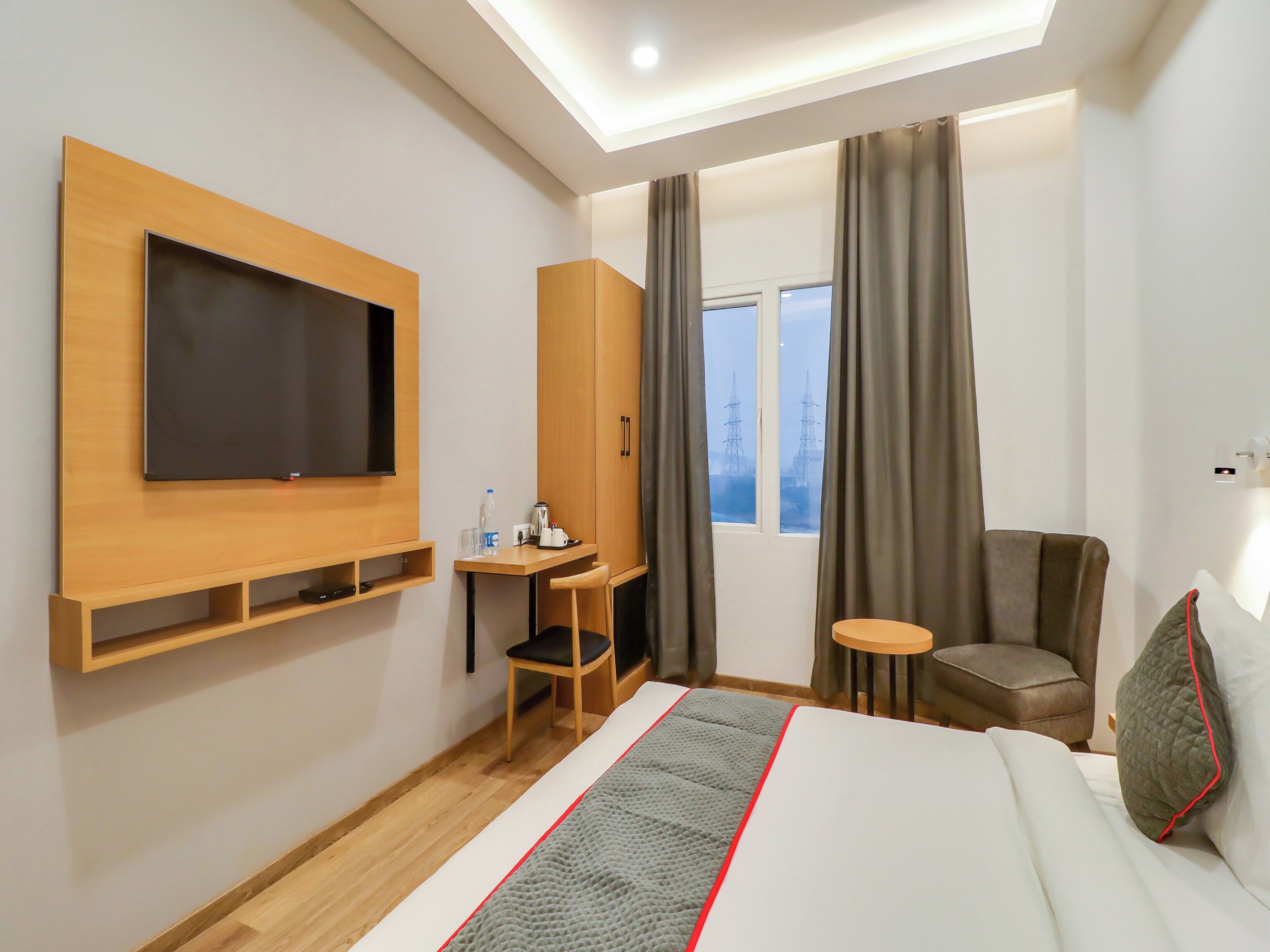 Una habitación de hotel con una cama, un televisor montado en la pared, un escritorio con una silla, una mesita y una ventana con cortinas.