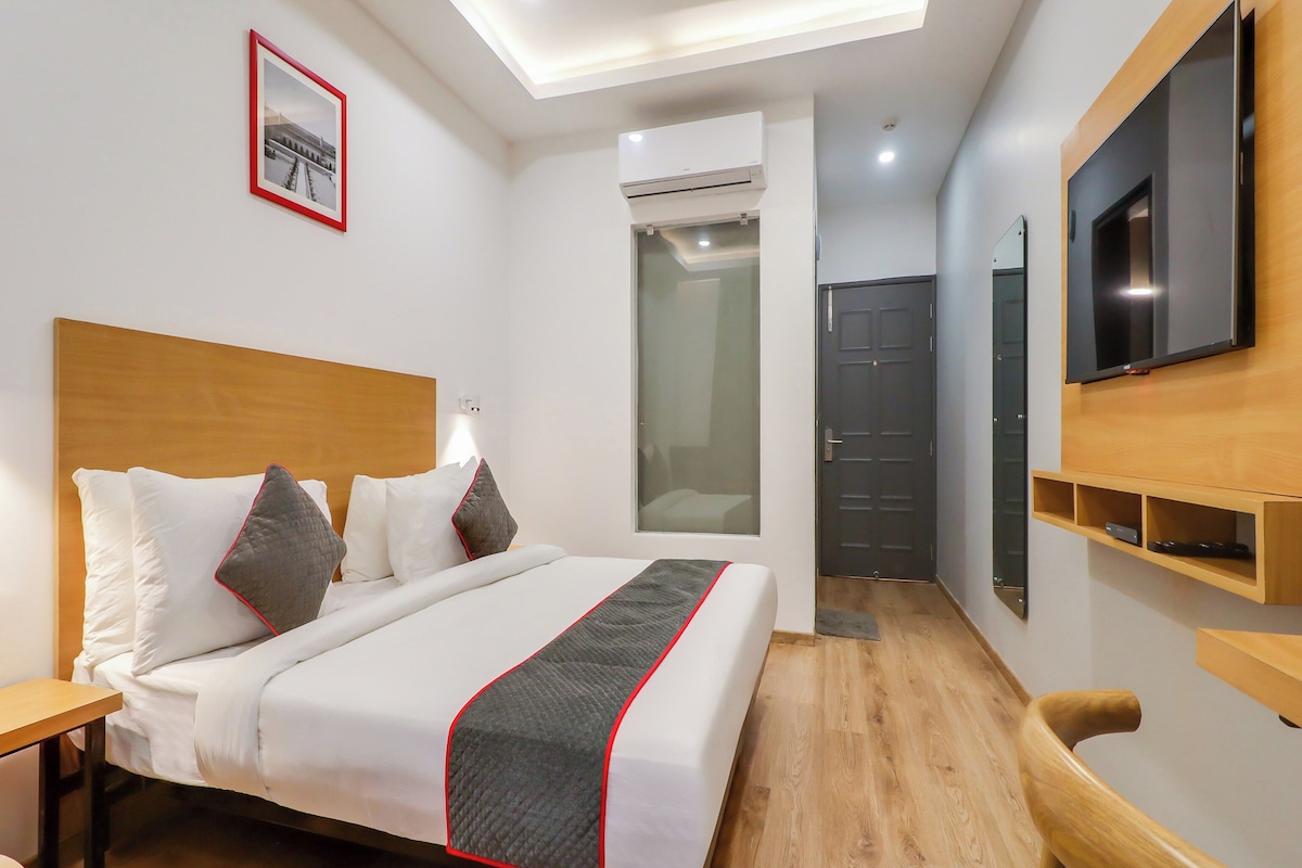 A hotel room with a bed, a TV, a wooden headboard, a bedside table, a wall-mounted shelf, and a door.
