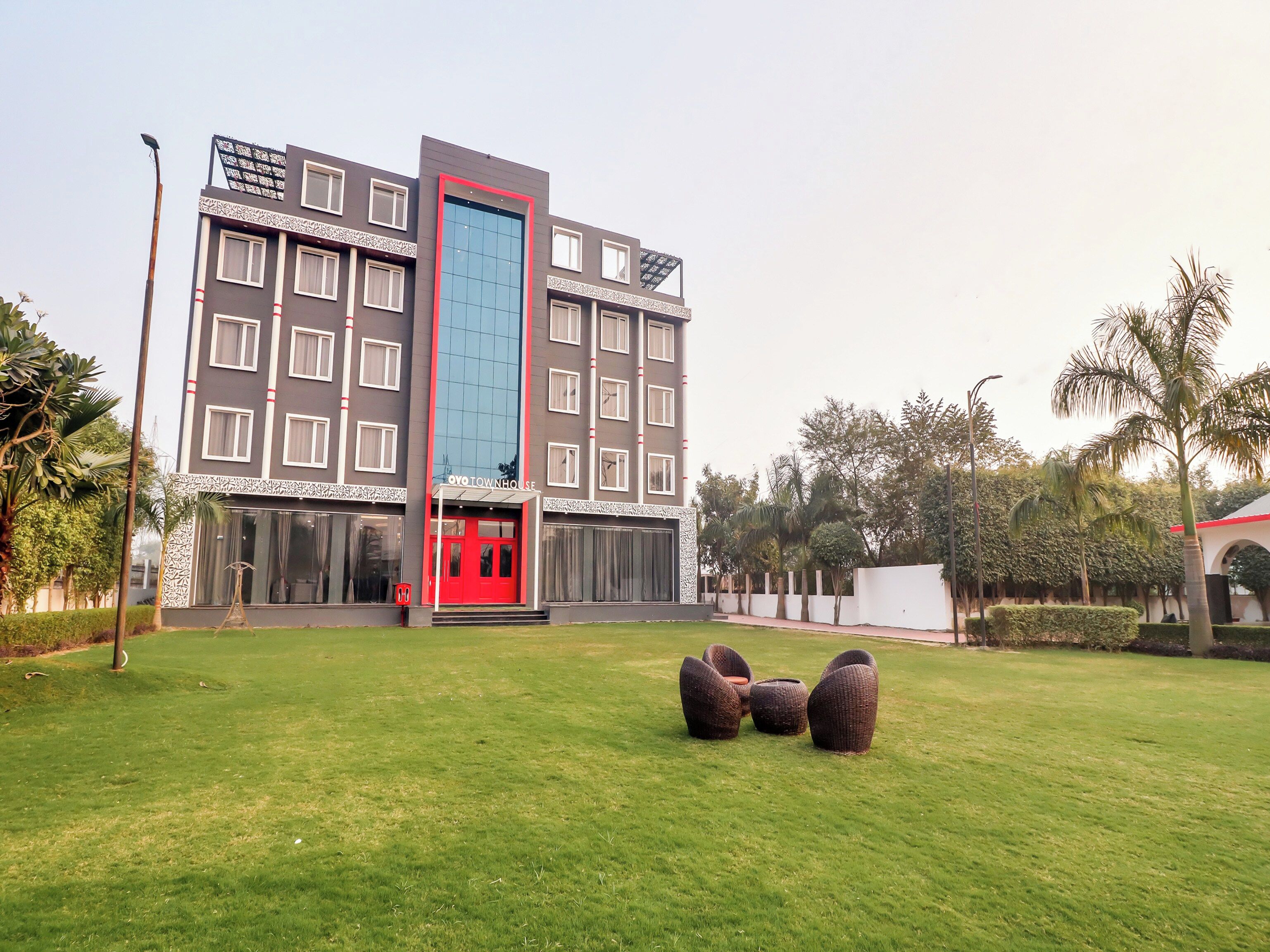 Un hotel con fachada moderna, entrada roja y césped cuidado con mobiliario exterior.