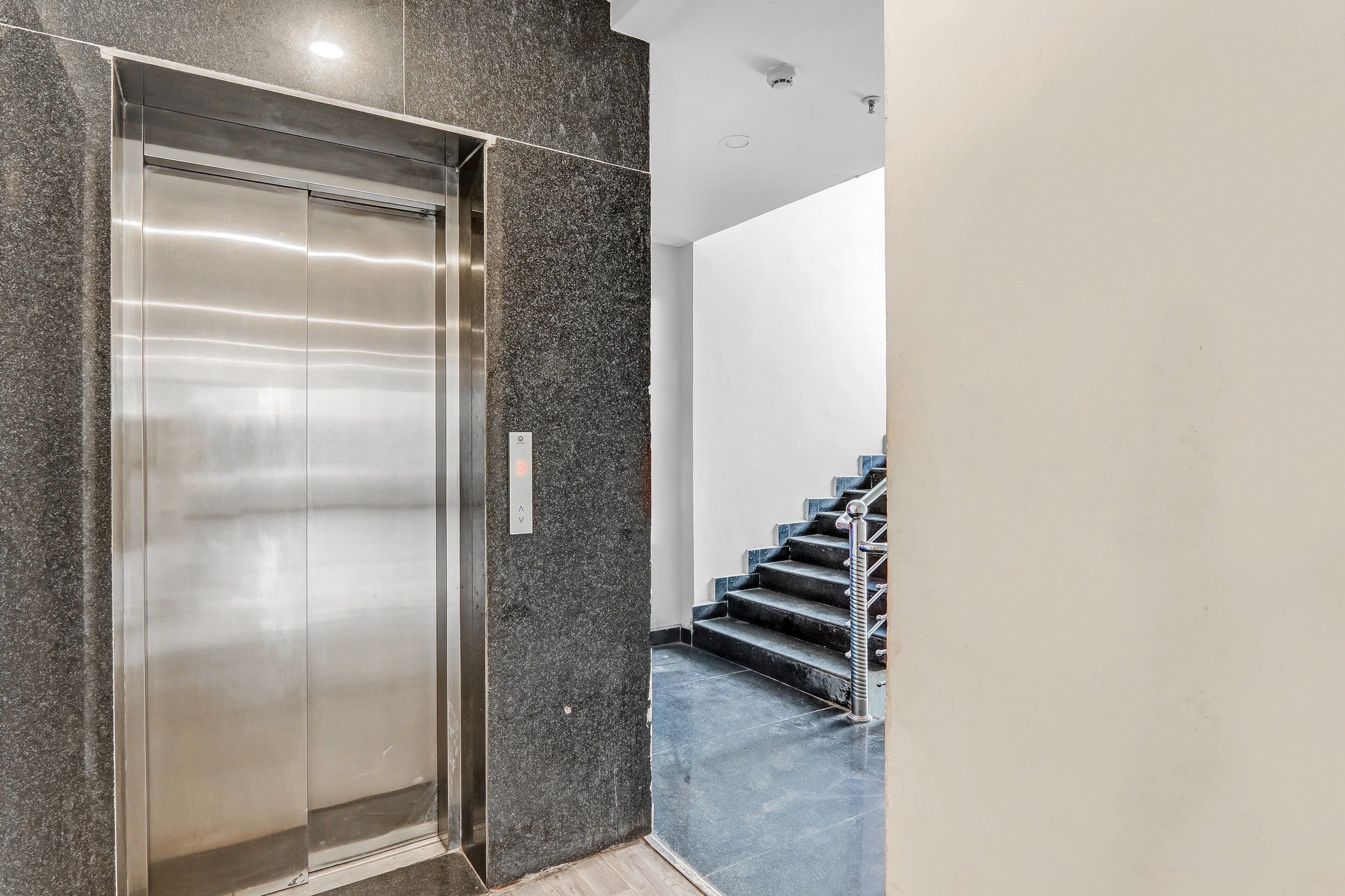 An elevator with a metallic finish, adjacent to a staircase with a metal handrail and dark steps.