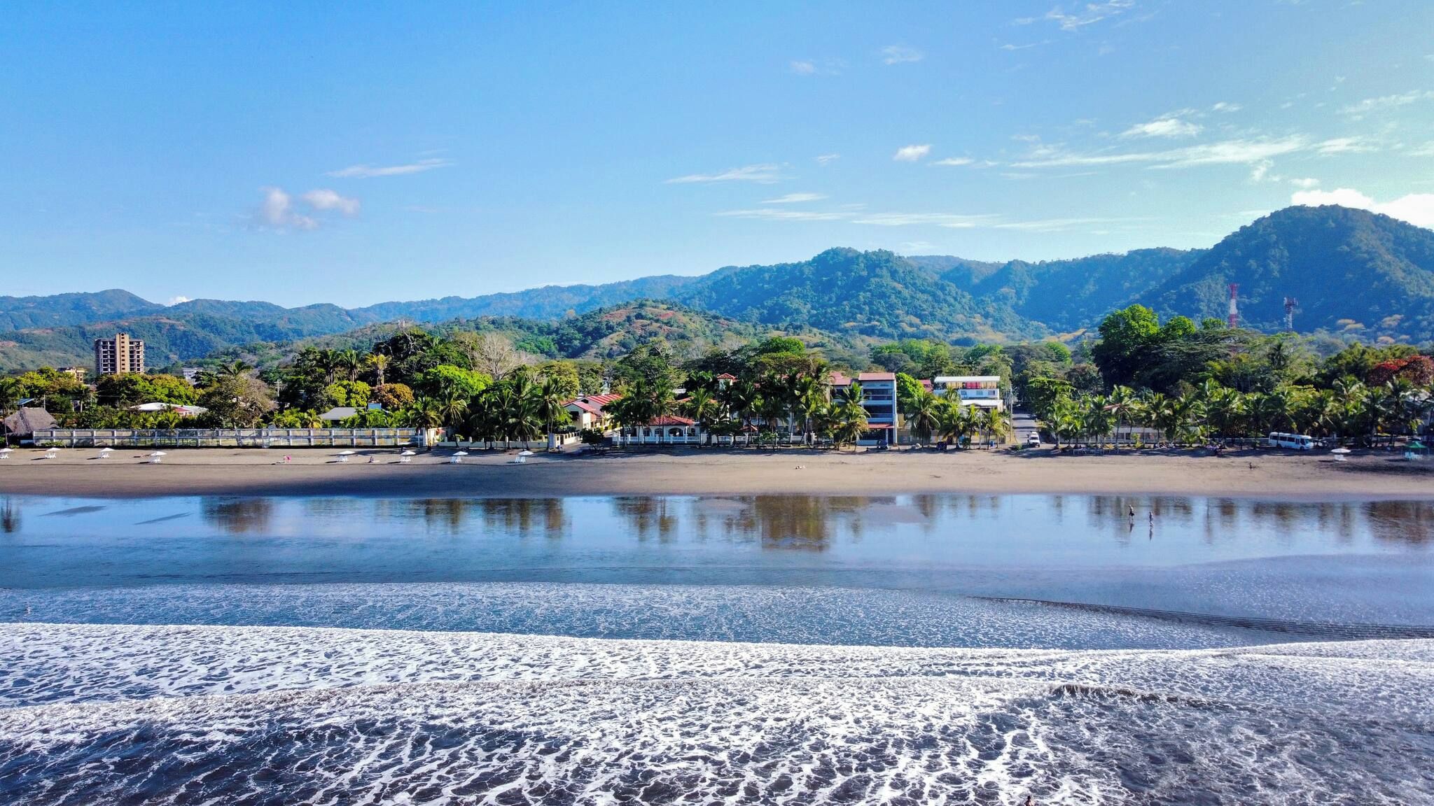 A beach with a calm sea, a sandy shore, and a backdrop of mountains and buildings.