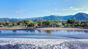 A beach with a calm sea, a sandy shore, and a backdrop of mountains and buildings. - Beachside Studio With Rooftop Views (Jacó)