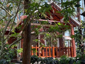 Une construction traditionnelle en bois, avec un toit rouge, entourée d’un feuillage dense et d’arbres.
